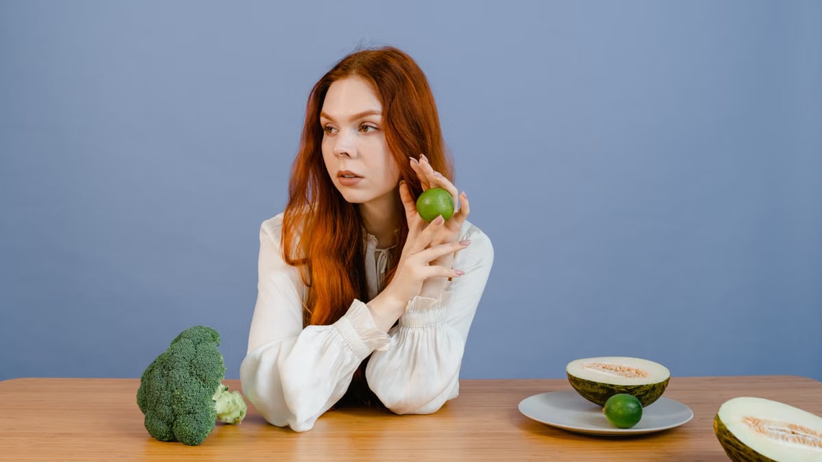 Caucasian woman sitting at table holding lime with broccoli and melon.